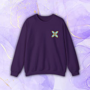 Könnte beinhalten: Ein lilafarbener Pullover mit einem kleinen, gestickten blauen und goldenen Schmetterlingsmotiv auf der linken Brust.