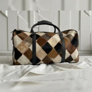Op de afbeelding: Een rechthoekige reistas met een patchwork-ontwerp. De tas heeft een patroon van bruine, zwarte en crèmekleurige ruitvormen. Hij heeft zwarte leren handvatten en riemen. De tas ligt op een witte doek.