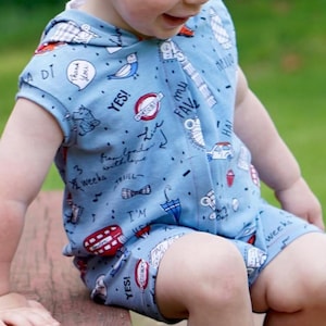 May include: A light blue hooded romper with a London theme, featuring illustrations of buses, umbrellas, and the text "YES!" and "I'M". The romper has short sleeves and legs, and is worn by a toddler.