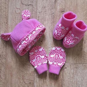Könnte beinhalten: Ein Set Babyaccessoires in leuchtendem Pink, bestehend aus einer Mütze mit ohrenartigen Ausbuchtungen, einem Paar Fäustlingen und einem Paar Babyschuhen. Jedes Teil hat ein weißes und rosa Schwanenmuster an den Rändern und auf den Schuhen.