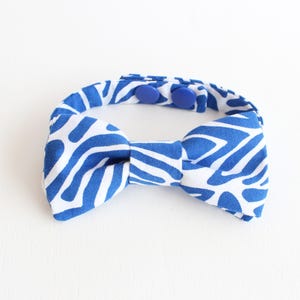 May include: A blue and white patterned bow tie with a matching adjustable collar. The bow tie features a bold, abstract design in blue against a white background. Two blue buttons secure the collar.