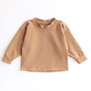 May include: A tan-coloured long-sleeved sweatshirt with a crew neck. The garment is made of a soft material and is laid flat on a white surface. The sleeves have a ribbed cuff.