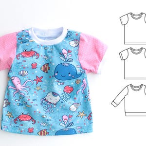 May include: A short-sleeved t-shirt featuring a whimsical ocean-themed print. The shirt has a light blue background with colorful illustrations of marine life, including whales, crabs, and jellyfish. The sleeves are pink with white polka dots, and the neckline is white.