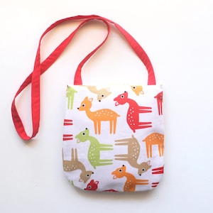 Puede incluir: Un bolso tote blanco con una correa roja presenta un colorido estampado de ciervos en naranja, verde y marr&oacute;n. Los ciervos est&aacute;n estilizados con formas simples y puntos. La bolsa est&aacute; dise&ntilde;ada para transportar objetos.
