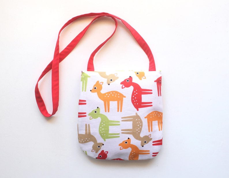 Puede incluir: Un bolso tote blanco con una correa roja presenta un colorido estampado de ciervos en naranja, verde y marr&oacute;n. Los ciervos est&aacute;n estilizados con formas simples y puntos. La bolsa est&aacute; dise&ntilde;ada para transportar objetos.