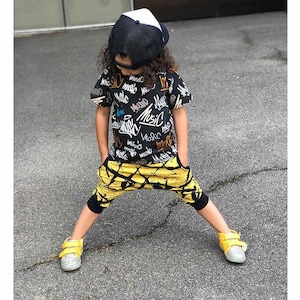 May include: A child wearing a black and white baseball cap, a black t-shirt with "MUSIC" graffiti, yellow and black patterned pants, and yellow sneakers. The child is standing with their legs apart.