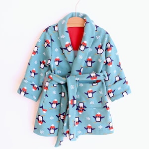 May include: A light blue children's robe with a penguin pattern. The robe has a red interior, a matching belt, and a shawl collar. The penguins are wearing red hats and are surrounded by white snowflakes.