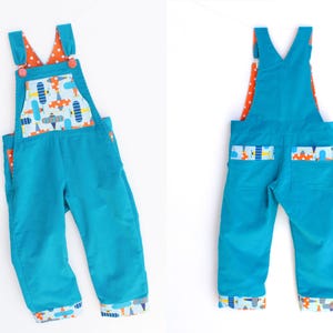 Puede incluir: Mono de pana turquesa con forro de lunares naranja y detalles de estampado de aviones. Cuenta con un bolsillo delantero y puños con un colorido estampado de aviones. Las correas se aseguran con botones rosas.