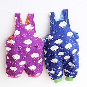 May include: Two pairs of baby overalls with a cloud and rain pattern. One pair is purple with orange lining and pink cuffs, the other is blue with a light blue lining and green cuffs. Both have white cloud graphics and colorful rain.