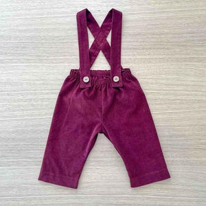 May include: Plum-colored corduroy overalls with suspenders. The suspenders cross in the back and attach to the waistband with wooden buttons. The pants have a relaxed fit and are designed for a child.