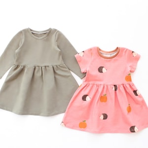 May include: Two children's dresses. One is a solid sage green with long sleeves. The other is pink with a pattern of hedgehogs and orange apples. Both dresses have a round neckline and a gathered skirt.