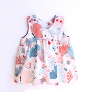 May include: A sleeveless dress in white, patterned with coral, blue, and red floral designs. It has a round neckline and red buttons on the shoulders. The lightweight fabric makes it suitable for warmer weather. The dress is approximately 60 cm in length.