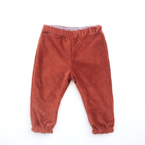 Puede incluir: Pantalones de bebé color óxido con cintura elástica y tobillos fruncidos. Los pantalones están hechos de un material suave, similar a la pana. El fondo es blanco liso.