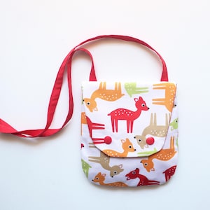 Puede incluir: Un peque&ntilde;o bolso cruzado blanco con una correa roja. El bolso presenta un patr&oacute;n repetido de ciervos coloridos en tonos de rojo, naranja, verde y marr&oacute;n. El cierre de solapa se asegura con botones rojos.