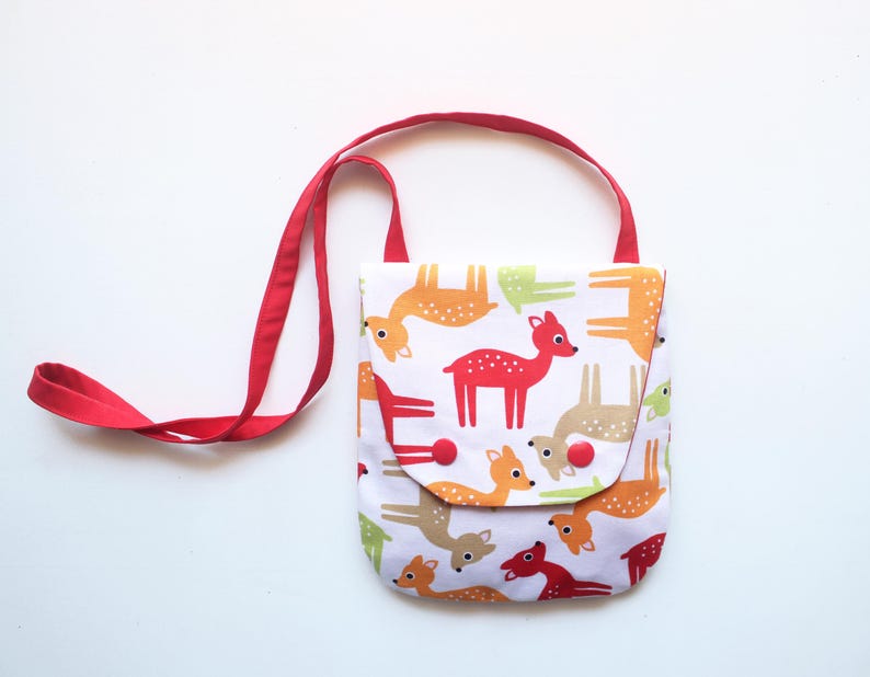 Puede incluir: Un peque&ntilde;o bolso cruzado blanco con una correa roja. El bolso presenta un patr&oacute;n repetido de ciervos coloridos en tonos de rojo, naranja, verde y marr&oacute;n. El cierre de solapa se asegura con botones rojos.
