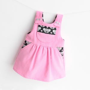 Op de afbeelding: Een roze baby tuinbroekjurk met een koala-berenpatroon op de zak en zijpanelen. De jurk heeft verstelbare bandjes met knopen en een gerimpelde rok. Het koalapatroon bevat grijze koala's en witte hartjes op een zwarte achtergrond.