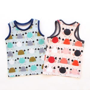 Puede incluir: Dos camisetas sin mangas con estampados de cangrejos. Una es azul claro con ribete azul marino y presenta cangrejos marrones, grises y turquesas. La otra es rosa con ribete gris y tiene cangrejos rojos, negros y rosas. Sobre fondo blanco.