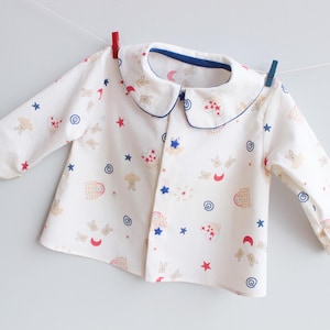 Pode incluir: Uma camisa de bebê branca com gola Peter Pan e mangas compridas, decorada com um padrão de coelhos, estrelas e outras formas em vermelho, azul e bege. A gola e os punhos são adornados em azul.
