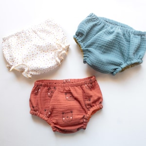 Op de afbeelding: Drie babybloomers in verschillende kleuren en patronen. Eén paar is wit met kleine, kleurrijke stippen en ruches. Een ander is lichtblauw en de derde is rood-oranje met berengezichten.