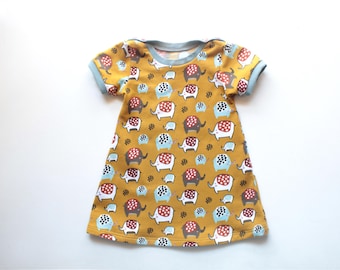 May include: Mustard yellow baby dress with a pattern of elephants in red, white, blue, and brown. The dress has short sleeves and a light blue trim around the neckline and sleeves. The dress is made of soft fabric.