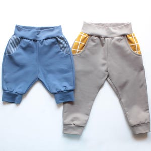 Puede incluir: Dos pantalones de bebé. Un par es azul claro con detalles de bolsillo a rayas. El otro par es gris con detalles de bolsillo a cuadros amarillos. Ambos tienen cinturillas y puños elásticos.