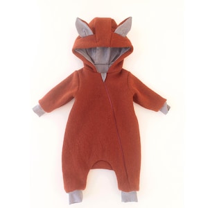 Könnte beinhalten: Ein rostfarbener Baby-Overall mit Kapuze und grauen Fuchsohren. Der Overall hat lange Ärmel und Bündchen, mit einem grauen Futter, das in der Kapuze und an den Bündchen sichtbar ist. Das Outfit ist so konzipiert, dass es den gesamten Körper bedeckt.