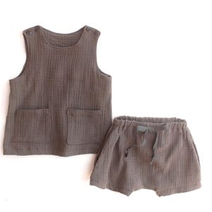 Könnte beinhalten: Ein zweiteiliges Baby-Outfit in einem gedämpften Braunton. Das Set besteht aus einem ärmellosen Oberteil mit zwei Vordertaschen und Knopfverschlüssen an den Schultern sowie passenden Shorts mit Kordelzug. Der Stoff hat ein strukturiertes, geripptes Aussehen.
