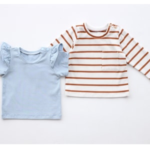 May include: Two baby shirts. One is light blue with ruffled sleeves. The other is a long-sleeved shirt with brown and white horizontal stripes and a small pocket on the front. Both shirts have a round neckline.