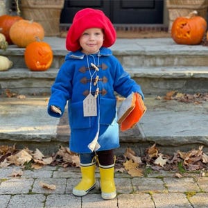 Puede incluir: Un niño vestido con un abrigo azul con cierres de madera y un sombrero rojo, sosteniendo un sándwich. Botas de agua amarillas y calabazas completan el atuendo otoñal.