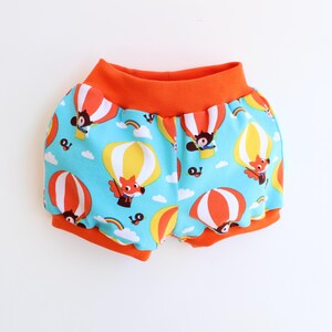 Puede incluir: Shorts de bebé con un estampado caprichoso de globos aerostáticos. Los pantalones cortos tienen un fondo azul claro con globos aerostáticos naranjas, amarillos y blancos y animales de dibujos animados. La cintura y las aberturas de las piernas son naranjas.