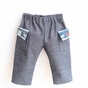 May include: Gray denim pants with two front pockets. Each pocket has a colorful patterned accent with a castle and insect design. The pants have an elastic waistband and are designed for children.