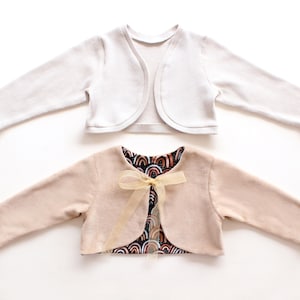 Könnte beinhalten: Zwei Kinder-Bolero-Sweater. Einer ist weiß, der andere ist hellbraun mit einem Regenbogenmuster-Futter und einer goldenen Schleife. Beide Pullover haben lange Ärmel und eine abgerundete Front.
