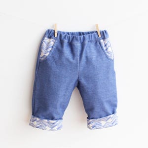 Op de afbeelding: Blauwe babybroek met gedetailleerde zakken en manchetten met patroon, hangend aan een waslijn. De broek is gemaakt van een zachte stof en heeft een comfortabele elastische tailleband. De manchetten en zakken hebben een wit en blauw zigzagpatroon.