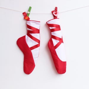 May include: Two ballet slipper-shaped Christmas stockings. The stockings are red and white with red ribbon accents. One is clipped with a green clip and the other with a pink heart clip, hanging from a white string.