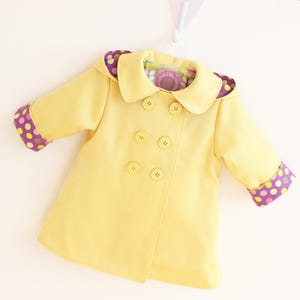 May include: A yellow double-breasted coat with a rounded collar and four buttons. The cuffs and hood lining feature a purple polka dot pattern. The coat is designed for a child.