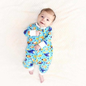 Può includere: Un bambino indossa una tutina blu a maniche lunghe con un motivo di pesci colorati. La tutina ha polsini e finiture bianche. Il bambino è sdraiato su una superficie bianca e strutturata.