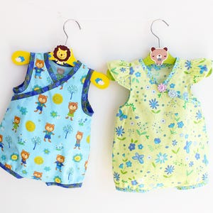 May include: Two baby rompers hanging on decorative hangers. One is light blue with a bear and tree pattern, the other is yellow with a floral design. Both have a wrap-style front and are sleeveless.