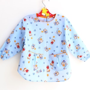 May include: A light blue, long-sleeved child's art smock with a round neckline and two front pockets. The smock is patterned with cartoon bears, hearts, and flowers. The smock is hanging on a yellow giraffe-shaped hanger.