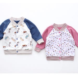 May include: Two children's bomber jackets. One has blue sleeves and a white body with a car pattern. The other has pink sleeves and a white body with a polka dot pattern. Both have button closures and ribbed cuffs.