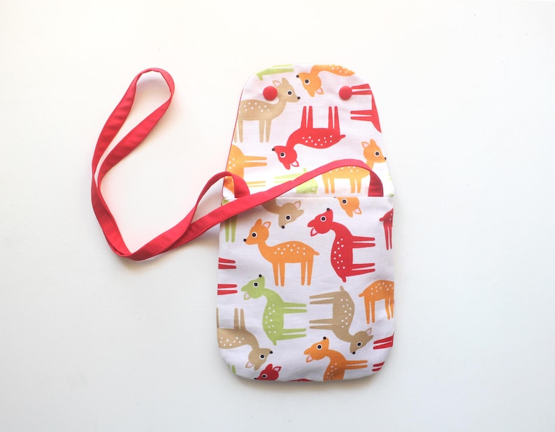 Puede incluir: Un bolso cruzado de tela blanca con una correa roja. El bolso tiene un estampado de ciervos coloridos en naranja, verde y rojo. El bolso tiene un cierre de solapa con un bot&oacute;n rojo.