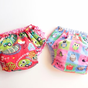 Puede incluir: Dos pañales de tela para bebés con diseños coloridos. Uno es rojo con un patrón caprichoso de coches, árboles y otras formas. El otro es un mosaico de cuadrados con búhos y otros animales en rosa, azul y verde.