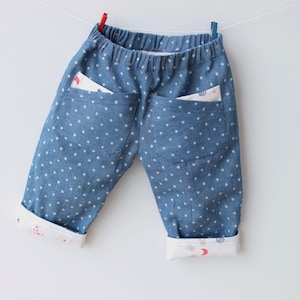 Puede incluir: Pantalones de bebé de mezclilla azul con estampado de estrellas blancas. Los pantalones tienen dos bolsillos traseros con tela estampada a la vista. Los puños están doblados, revelando un forro estampado. Los pantalones cuelgan de un tendedero con pinzas rojas y azules.