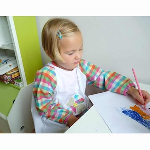 May include: A young child wearing a white art smock with colorful plaid sleeves is drawing on white paper with a pink pencil. The smock has a floral design on the front. The child is sitting at a white table.