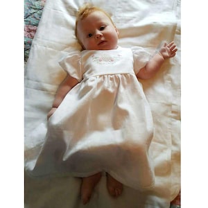 May include: A baby wearing a white dress with short sleeves and a floral embroidery on the bodice. The baby is lying on a white blanket. The dress is a classic style, suitable for a christening or special occasion.