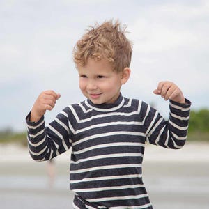 Puede incluir: Un niño con una camisa de manga larga a rayas azul marino y blanco con cuello alto. La camisa tiene rayas horizontales y está hecha de un material suave. El niño sonríe y flexiona los brazos.