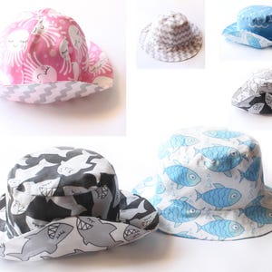 May include: A collection of reversible bucket hats in various patterns. One hat features a pink background with white smiling suns and octopus designs, while another displays a black and white shark pattern. Other hats have blue fish, gray and white chevron, and blue wave patterns.