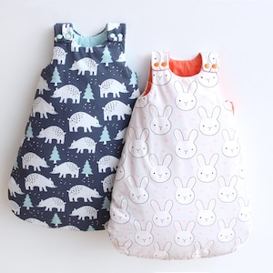 May include: Two baby sleep sacks. One is navy blue with white bear and tree patterns. The other is light pink with white bunny faces and small pink hearts. Both have button closures.