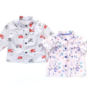 Può includere: Due camicie abbottonate per bambini. Una camicia bianca a maniche lunghe con un motivo di veicoli rossi e grigi. L'altra è una camicia a maniche corte con un motivo di mulini a vento e uccelli. Entrambe hanno il colletto.