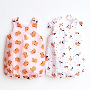 May include: Two baby rompers. One is light pink with orange tiger faces. The other is white with orange fruit and black dots. Both have snap closures and tie straps.
