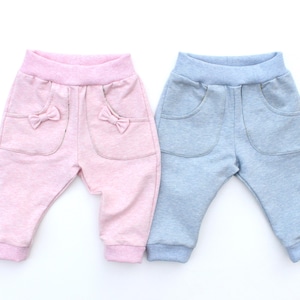 Può includere: Due pantaloni per neonati in colori pastello. Uno è rosa chiaro con fiocchi sulle tasche, l'altro è azzurro. Entrambi hanno una comoda fascia elastica in vita e polsini.